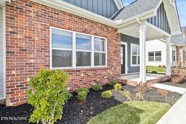 doorway to property with brick siding, board and batten siding, a porch, and roof with shingles