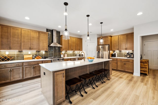 kitchen with wall chimney range hood, a kitchen breakfast bar, appliances with stainless steel finishes, light wood-style floors, and brown cabinetry