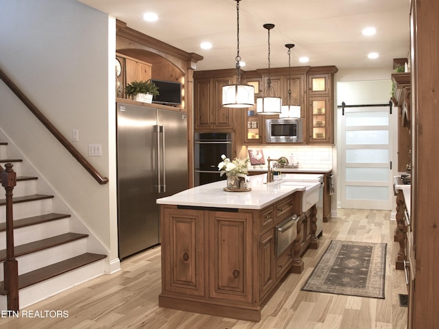 kitchen with an island with sink, backsplash, stainless steel appliances, glass insert cabinets, and hanging light fixtures