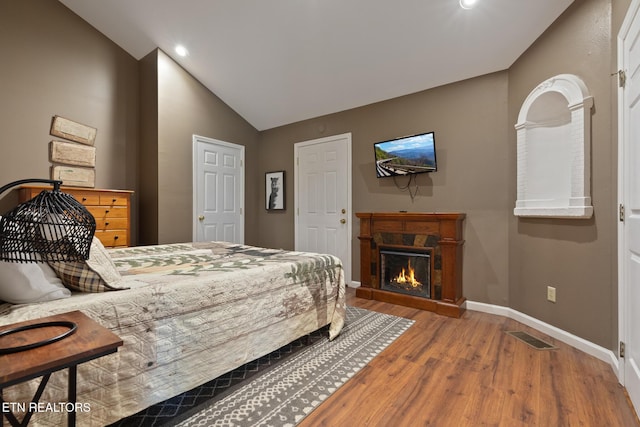 bedroom with visible vents, baseboards, lofted ceiling, a warm lit fireplace, and wood finished floors