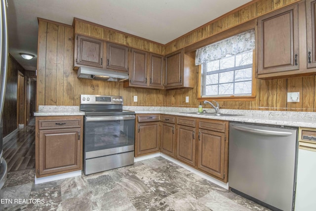 kitchen featuring wood walls, appliances with stainless steel finishes, under cabinet range hood, a sink, and brown cabinetry