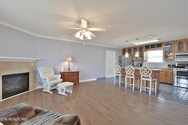 living area with a ceiling fan, a tile fireplace, baseboards, ornamental molding, and dark wood-type flooring