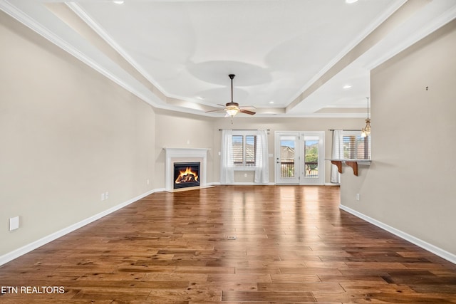 unfurnished living room with a tray ceiling, a fireplace with flush hearth, baseboards, a ceiling fan, and wood finished floors
