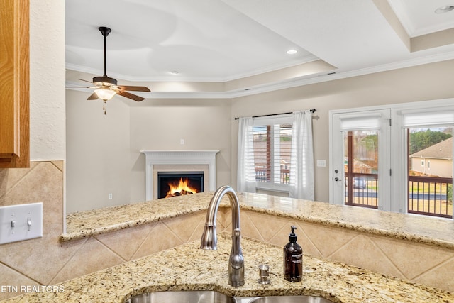 kitchen with ceiling fan, ornamental molding, a lit fireplace, a sink, and a tray ceiling
