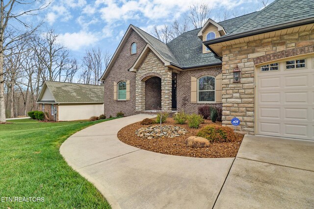 french country home with stone siding, roof with shingles, brick siding, a front lawn, and a garage