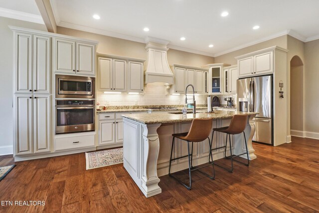 kitchen featuring an island with sink, light stone counters, appliances with stainless steel finishes, premium range hood, and arched walkways