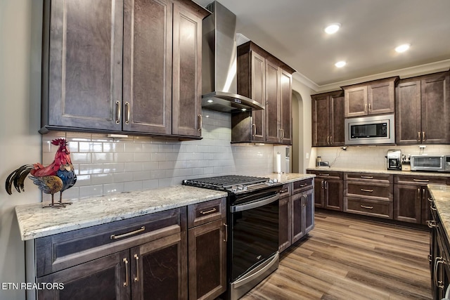 kitchen with wood finished floors, stainless steel microwave, dark brown cabinets, wall chimney range hood, and gas range oven