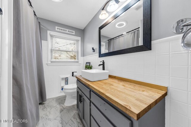 full bath with toilet, vanity, wainscoting, marble finish floor, and tile walls
