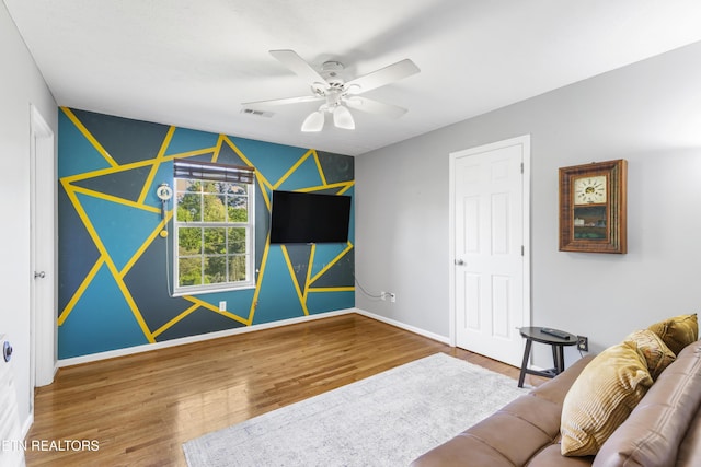 living room with a ceiling fan, visible vents, baseboards, an accent wall, and wood finished floors
