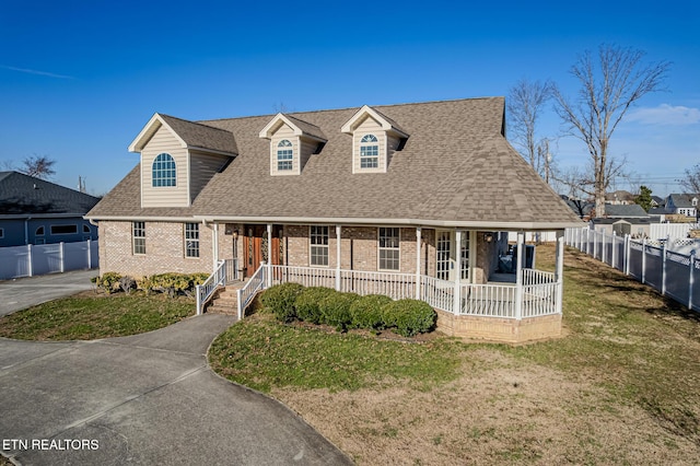 cape cod home with a front yard, covered porch, roof with shingles, fence, and brick siding