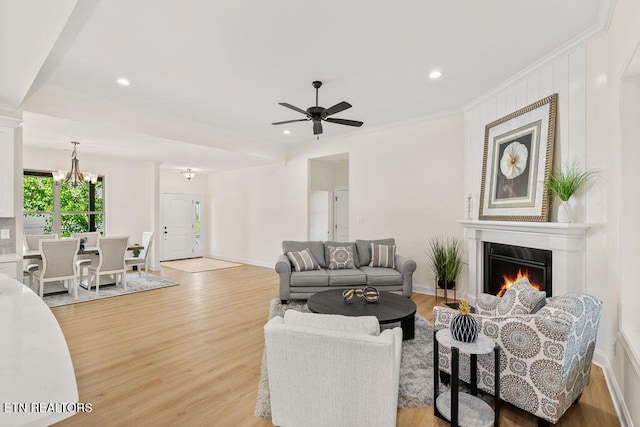 living room with recessed lighting, light wood-style floors, ornamental molding, ceiling fan with notable chandelier, and a lit fireplace