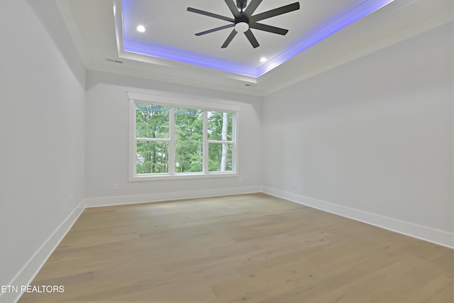 spare room featuring a raised ceiling, a ceiling fan, baseboards, crown molding, and light wood-style flooring