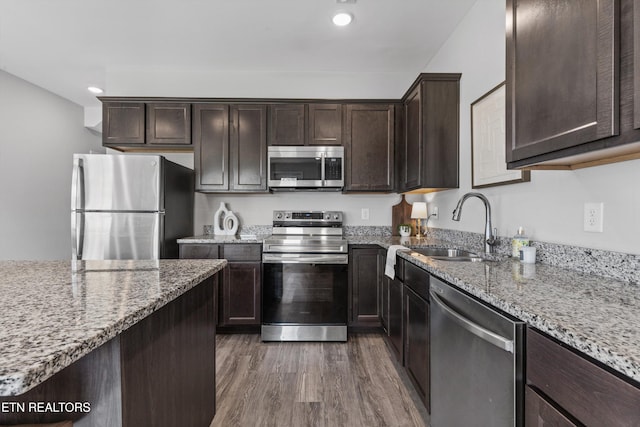 kitchen featuring light stone counters, dark brown cabinets, wood finished floors, a sink, and appliances with stainless steel finishes
