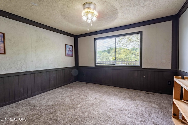 carpeted spare room featuring a ceiling fan, a textured ceiling, visible vents, wainscoting, and a textured wall
