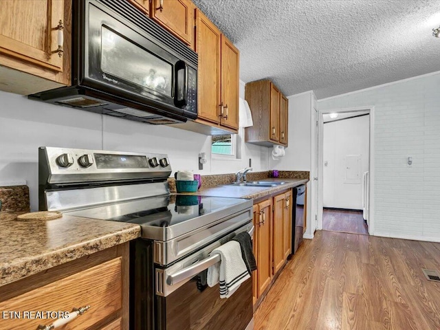 kitchen with a textured ceiling, black appliances, brown cabinetry, a sink, and wood finished floors