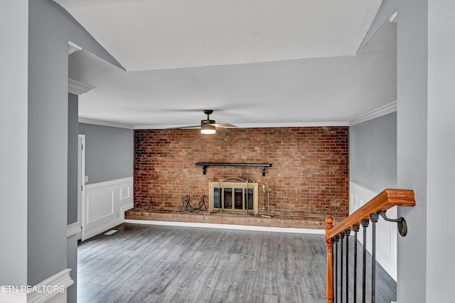unfurnished living room with ceiling fan, a fireplace, ornamental molding, a wainscoted wall, and wood finished floors