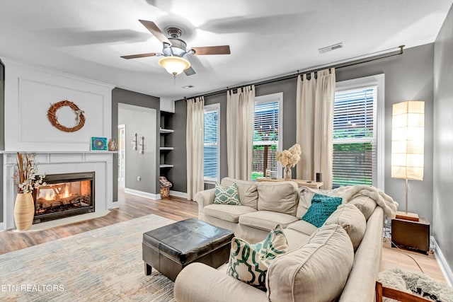 living area featuring built in features, a ceiling fan, visible vents, a warm lit fireplace, and wood finished floors