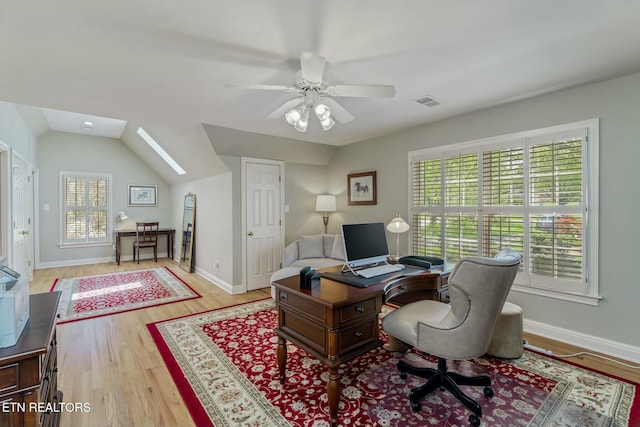 office area with baseboards, vaulted ceiling with skylight, light wood-style flooring, visible vents, and ceiling fan