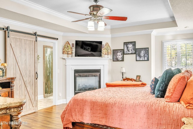 bedroom with a glass covered fireplace, light wood-type flooring, crown molding, a barn door, and a raised ceiling