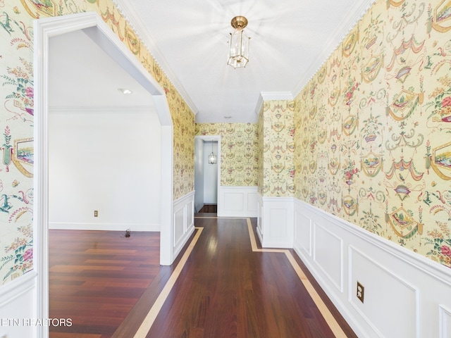 corridor with wallpapered walls, dark wood-style flooring, ornamental molding, and a wainscoted wall