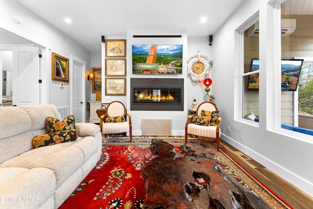 living area with a glass covered fireplace, baseboards, a wall unit AC, wood finished floors, and recessed lighting