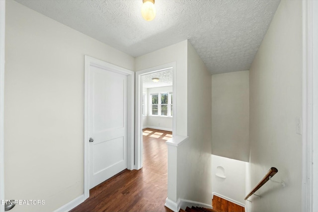 hall with a textured ceiling, baseboards, dark wood finished floors, and an upstairs landing