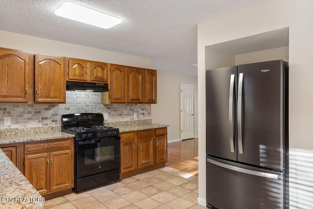 kitchen featuring freestanding refrigerator, tasteful backsplash, under cabinet range hood, brown cabinets, and black gas range oven