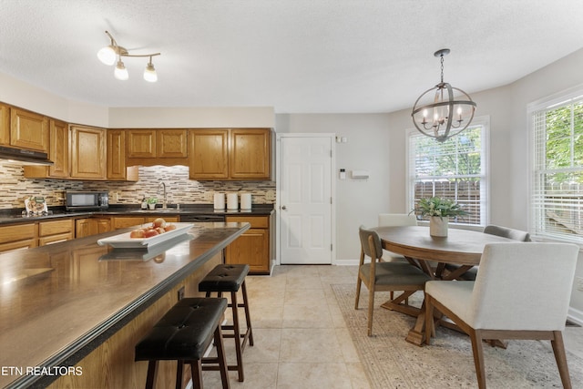 kitchen featuring brown cabinetry, backsplash, dark countertops, black appliances, and a notable chandelier