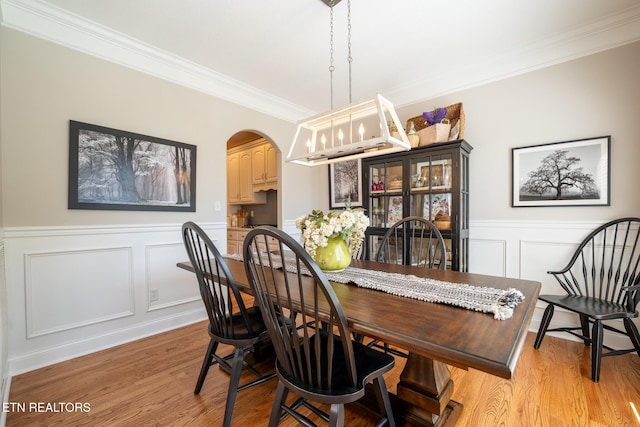 dining area featuring light wood-style flooring, a wainscoted wall, arched walkways, a decorative wall, and ornamental molding