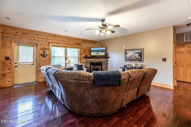 living area with a ceiling fan, a stone fireplace, baseboards, wooden walls, and dark wood-type flooring
