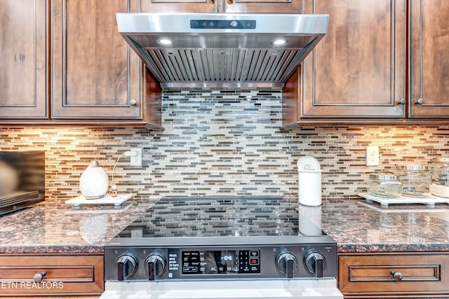 kitchen with stove, brown cabinetry, custom exhaust hood, dark stone countertops, and decorative backsplash