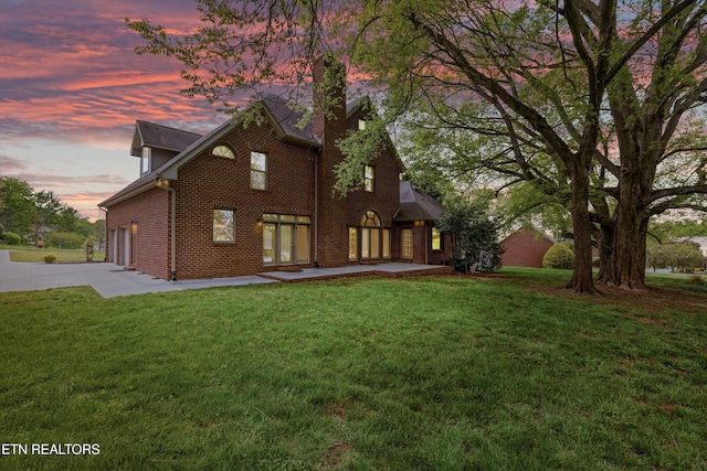 back of property featuring a chimney, concrete driveway, brick siding, a lawn, and a patio