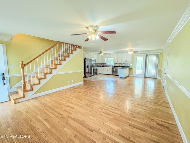 unfurnished living room featuring light wood finished floors, a ceiling fan, crown molding, baseboards, and stairs