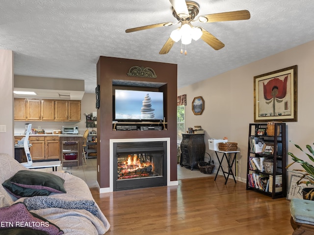 living room featuring a ceiling fan, a textured ceiling, wood finished floors, a toaster, and a warm lit fireplace