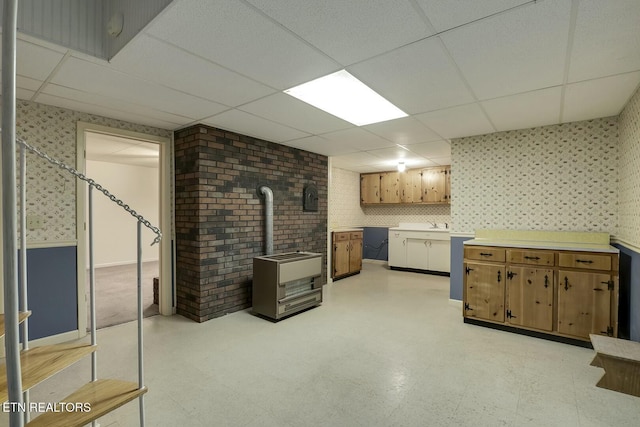 finished basement with wallpapered walls, a paneled ceiling, a wood stove, and light floors
