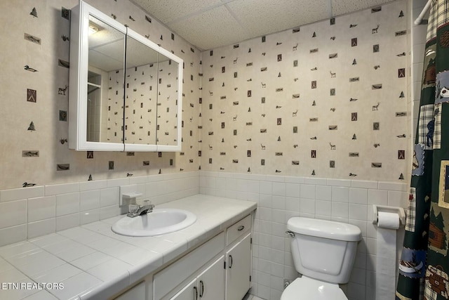 full bath featuring wallpapered walls, toilet, a wainscoted wall, and a drop ceiling