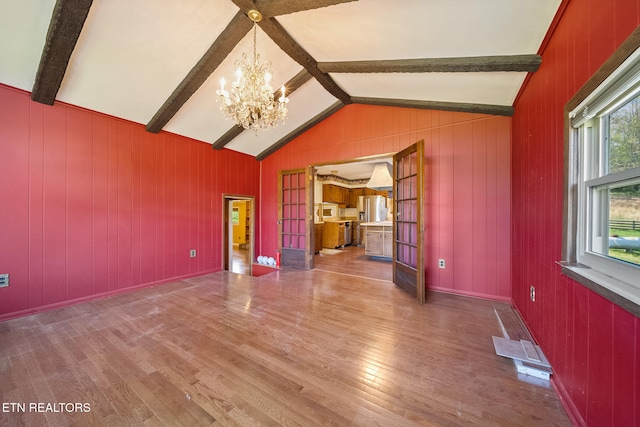 unfurnished living room featuring an inviting chandelier, lofted ceiling with beams, wooden walls, french doors, and wood finished floors
