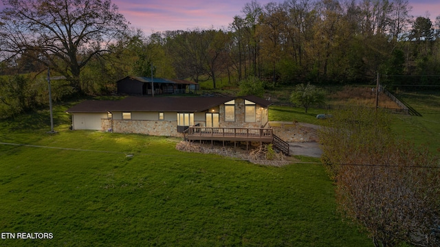 back of property at dusk featuring a wooded view, a deck, stone siding, and a lawn