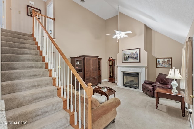 living area featuring high vaulted ceiling, stairs, a fireplace with flush hearth, ceiling fan, and light carpet
