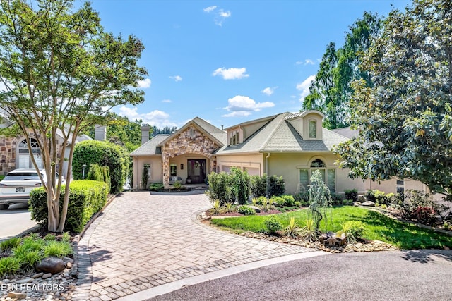 french country inspired facade featuring roof with shingles, a garage, decorative driveway, stone siding, and stucco siding