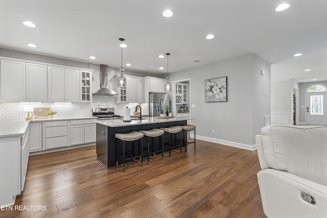 kitchen featuring glass insert cabinets, stainless steel appliances, open floor plan, wall chimney exhaust hood, and an island with sink