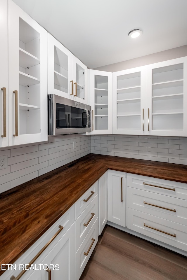 kitchen featuring stainless steel microwave, decorative backsplash, wood counters, glass insert cabinets, and white cabinetry