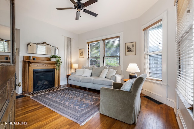 living area with dark wood finished floors, a glass covered fireplace, baseboards, visible vents, and a ceiling fan