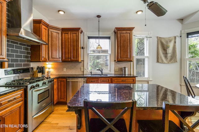 kitchen featuring wall chimney range hood, a kitchen breakfast bar, a sink, appliances with stainless steel finishes, and light wood-type flooring