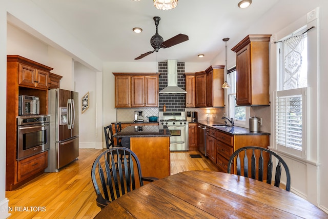 kitchen with wall chimney range hood, light wood-style flooring, appliances with stainless steel finishes, ceiling fan, and a kitchen island