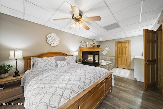 bedroom featuring a ceiling fan, a paneled ceiling, a multi sided fireplace, and dark wood-style floors
