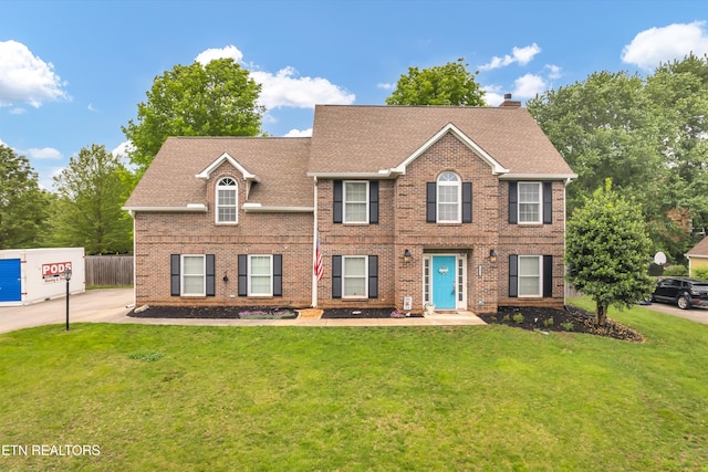 colonial-style house with a chimney, brick siding, fence, a shingled roof, and a front lawn