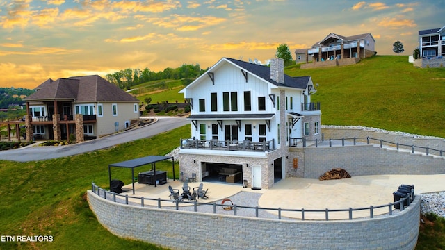 back of house at dusk with a chimney, an attached garage, a balcony, board and batten siding, and a yard