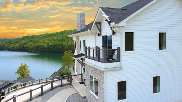 view of side of home with roof with shingles, a water view, a chimney, fence, and a balcony