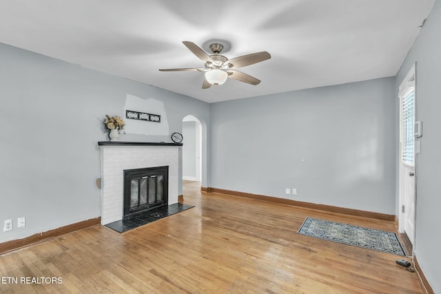 unfurnished living room featuring wood finished floors, a ceiling fan, arched walkways, baseboards, and a brick fireplace